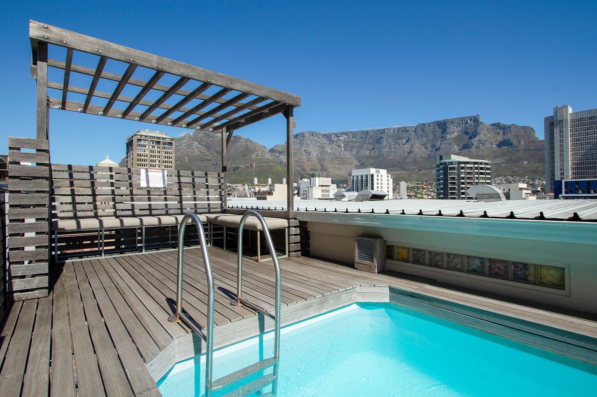 A rooftop pool with wooden deck, metal railings, and a pergola.