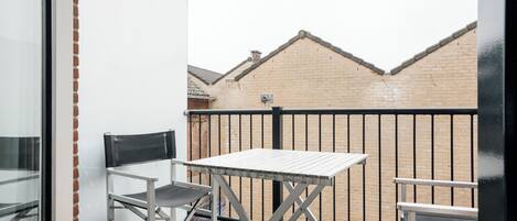 A balcony with a metal table and two chairs, a brick wall, and a metal railing in the background.