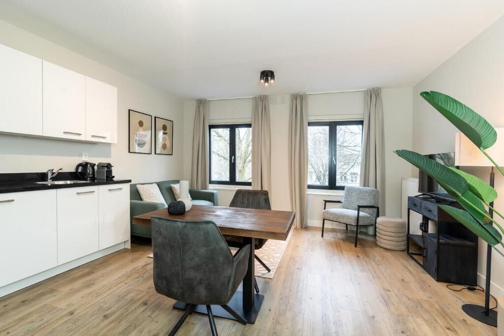 Una cocina moderna con armarios blancos, un comedor con mesa y sillas de madera, y ventanas grandes con cortinas.
