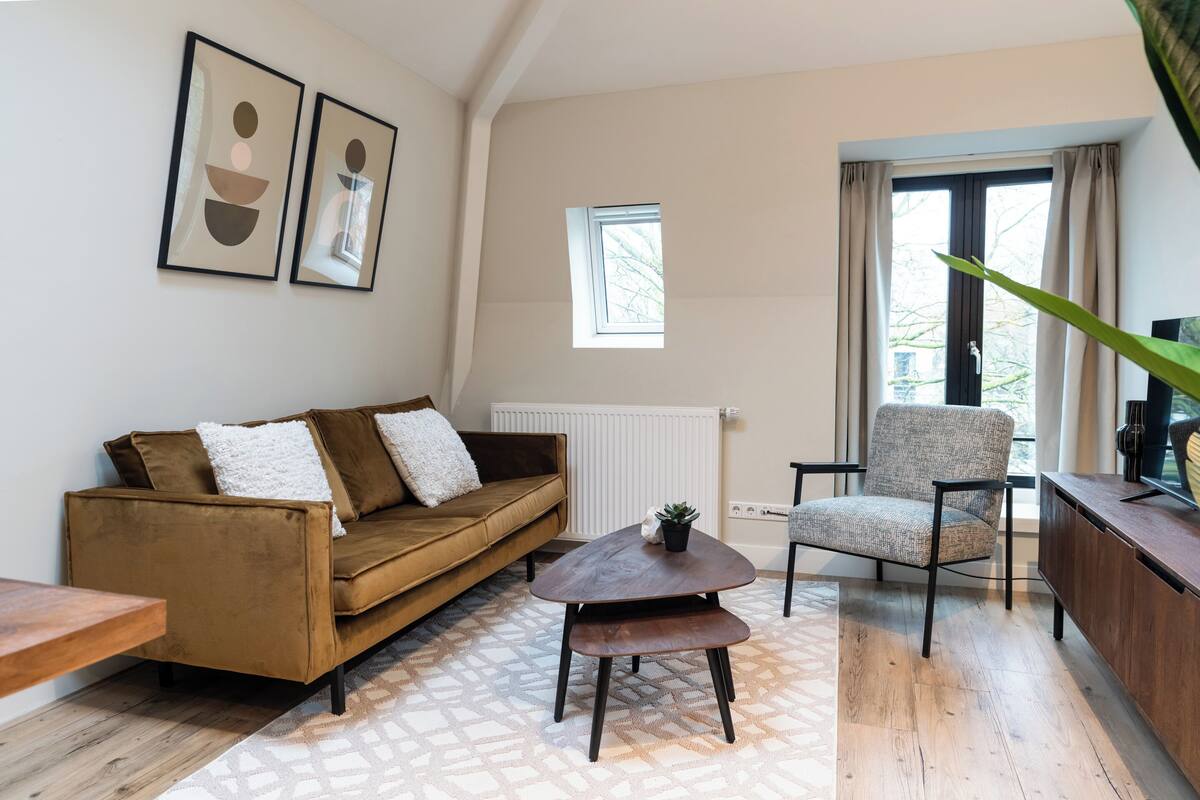 Ein Wohnzimmer mit einem braunen Sofa, einem Holztisch, einem gemusterten Teppich, einem grauen Sessel und einem Fenster mit Vorhängen.