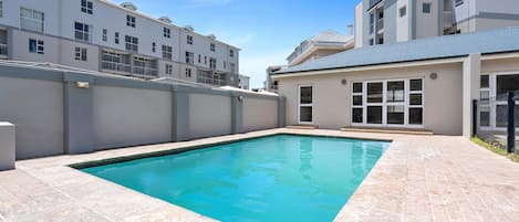 A swimming pool with a tiled deck, surrounded by a high wall and residential buildings.