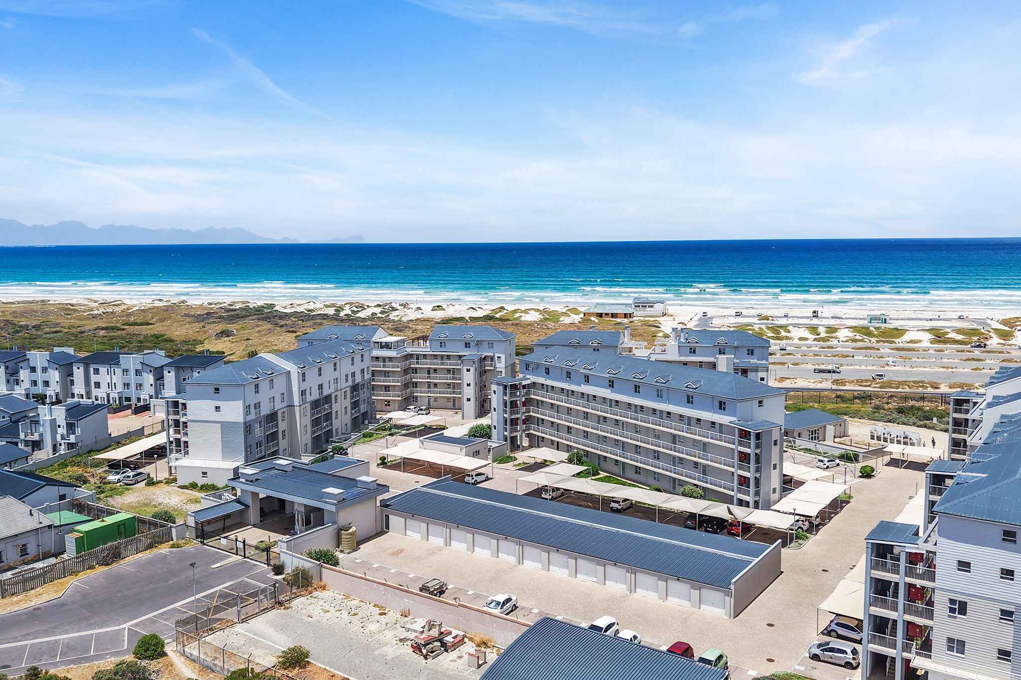 A coastal residential area with modern buildings, a parking lot, and a beach in the background.
