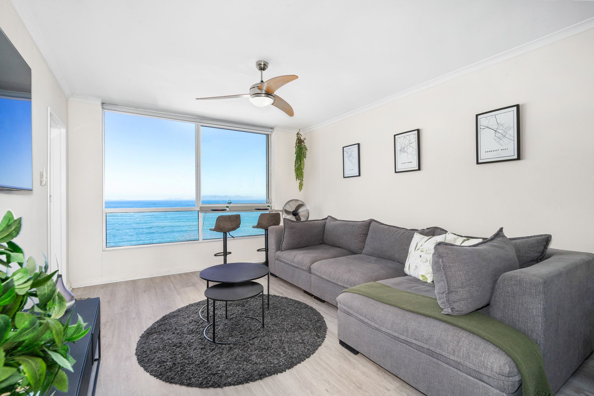 A modern living room with a large window overlooking the ocean, a gray sofa, a small table, and a ceiling fan.