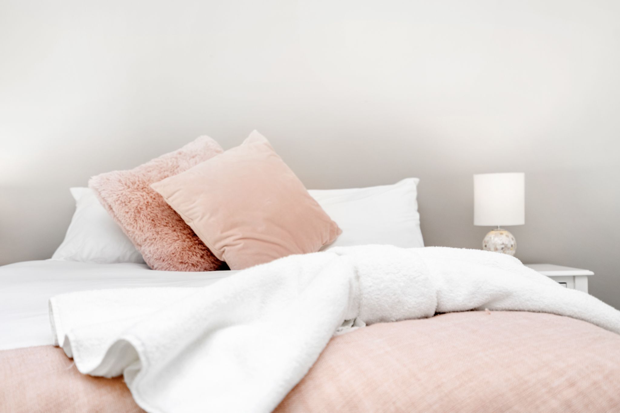 A neatly made bed with white and pink bedding, a bedside lamp, and a small white nightstand.