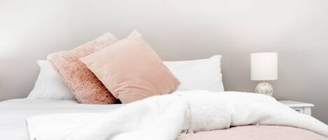 A neatly made bed with white and pink bedding, a bedside lamp, and a small white nightstand.