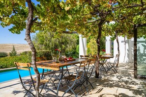 An outdoor dining area with a pool, wooden table, metal chairs, and a pergola with vines.