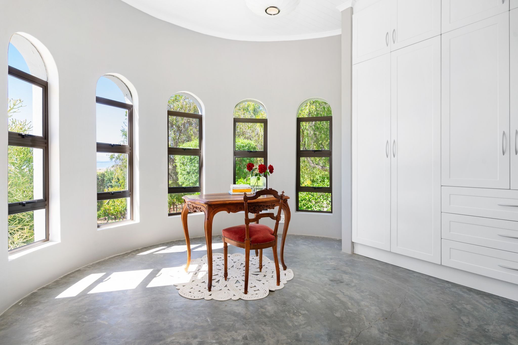 A room with a curved ceiling, white walls, and a large white cabinet. There are three arched windows with a view of greenery outside. A wooden table with a red chair and a vase of red flowers is placed in the center.
