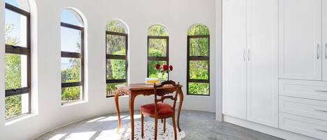 A room with a curved ceiling, white walls, and a large white cabinet. There are three arched windows with a view of greenery outside. A wooden table with a red chair and a vase of red flowers is placed in the center.