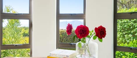 Une table en bois, ornée de livres et d’un vase de roses rouges, est placée près d’une fenêtre offrant une vue sur la verdure et les montagnes.