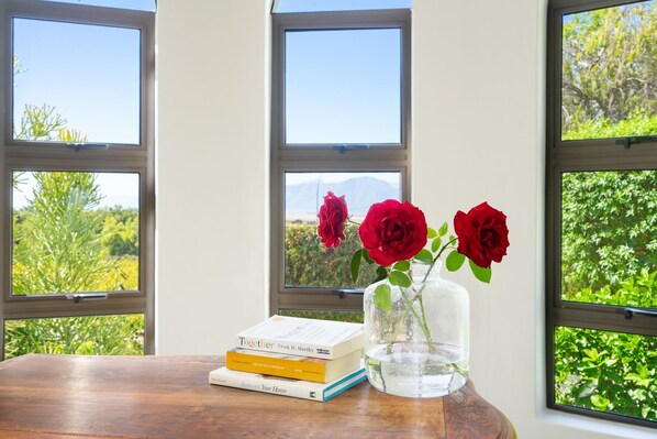 A wooden table with books and a vase of red roses by a window with a view of greenery and mountains.