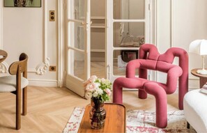 A modern living room with a wooden floor, a pink upholstered chair, a wooden coffee table with a vase of flowers, and a white sofa. - JUST MOMENTS FROM THE ICONIC CHAMPS-ÉLYSÉES & AVENUE MONTAIGNE - 2 BEDROOM (Paris)