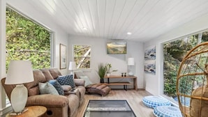 A modern living room with a sofa, coffee table, and decorative pillows. There are large windows with a view of greenery outside. - Luxury Cabin Retreat w/ Glass Floor Over Creek, 4BR/4BA Near Pigeon Forge, 5 AC (Sevierville)