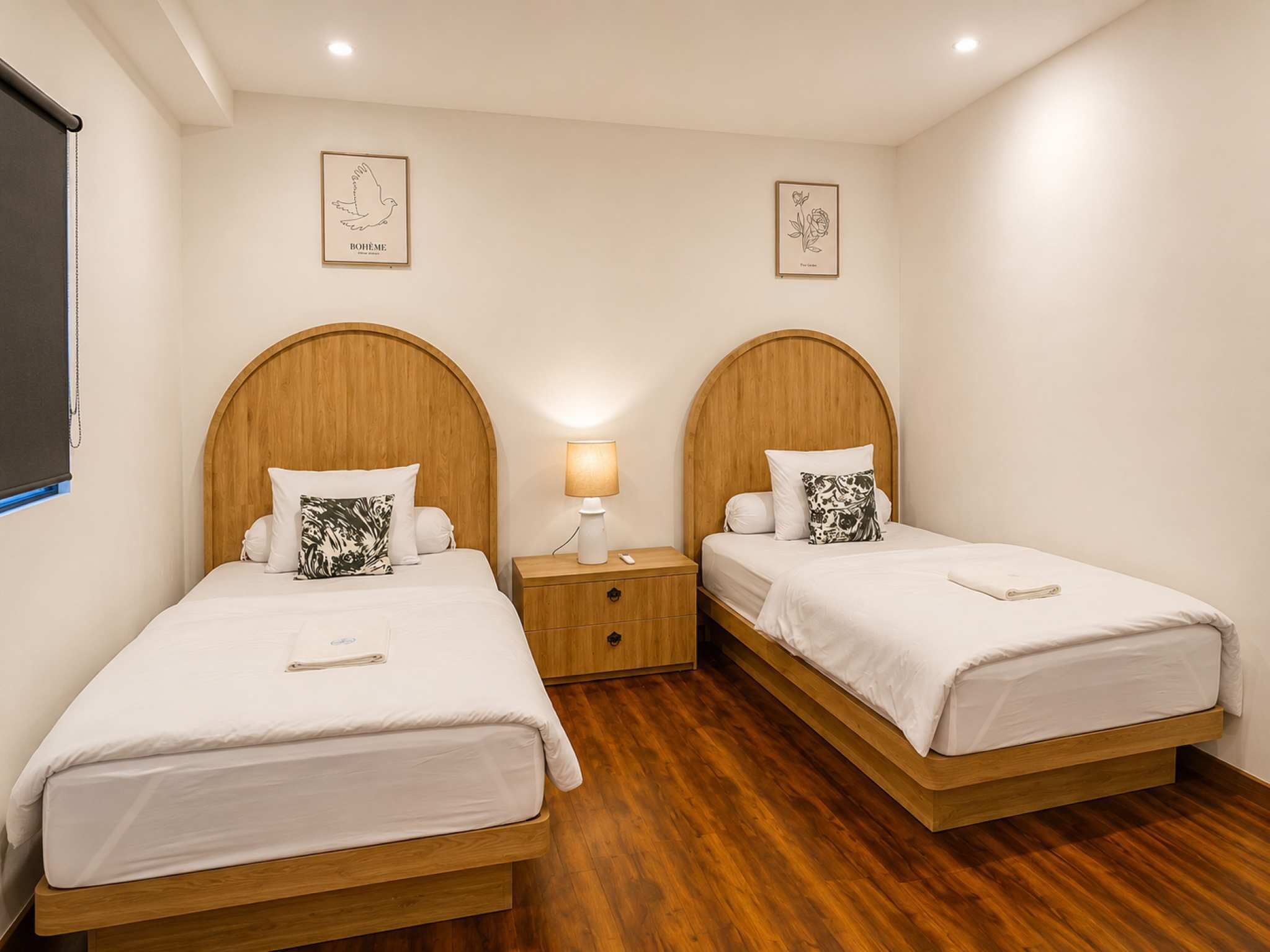 A double room with two wooden beds, a nightstand with a lamp, and two framed artworks on the walls.