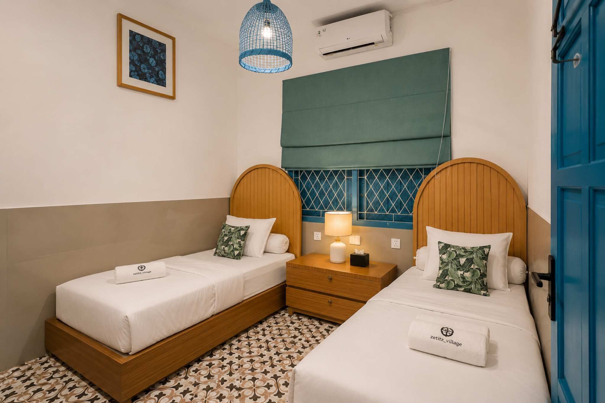 A hotel room with two beds, a nightstand, a lamp, a window with a green blind, and a blue door.