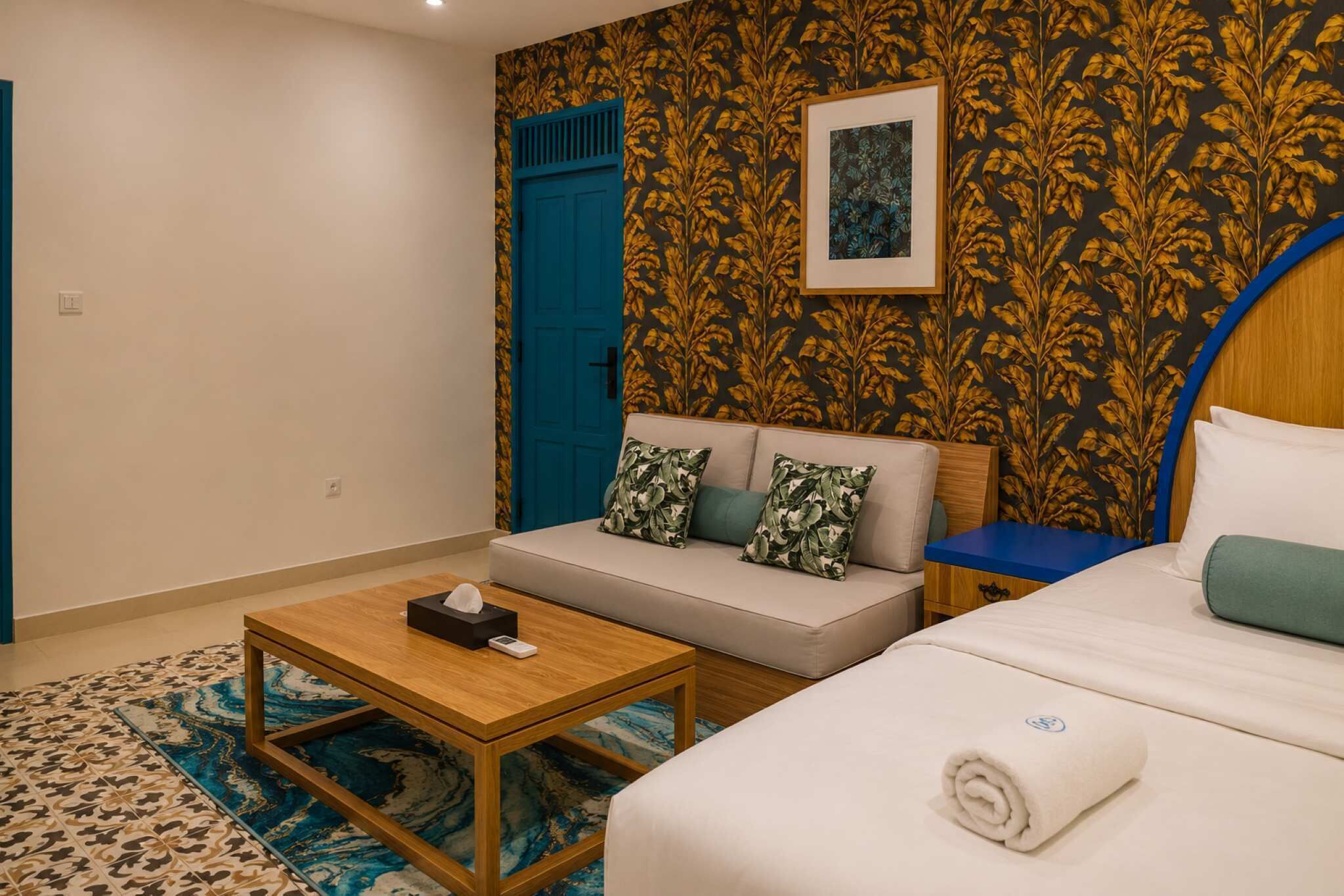 A hotel room with a sofa, a bed, a wooden table, a blue door, and a patterned wall.