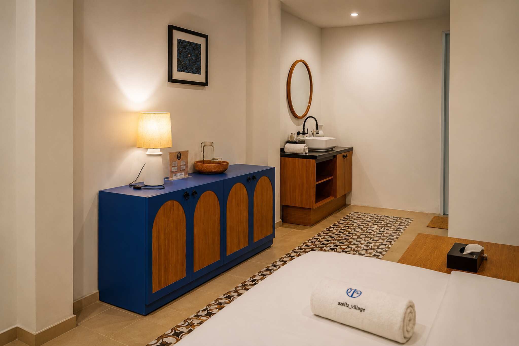A hotel room with a blue cabinet, a bedside table, a bed with a rolled towel, a small table with a lamp, a mirror, and a framed picture on the wall.