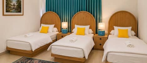 Quarto de hotel com três camas de solteiro, uma janela grande com cortinas azul-turquesa, uma luminária pendente azul e um tapete estampado.