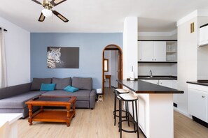 Ein modernes Wohnzimmer mit grauem Sofa, einem Holztisch und einer Küchenzeile mit weißen Schränken und einer schwarzen Arbeitsplatte.