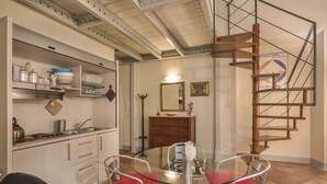 A compact kitchen with modern appliances, a dining area with a glass table and metal chairs, and a wooden spiral staircase.