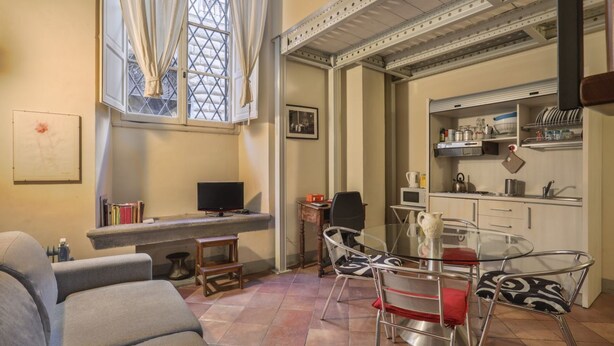 A compact living space with a sofa, a glass dining table, a kitchen area, and a window with curtains.