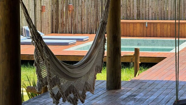 A hammock hanging between wooden posts, a wooden deck, and a pool area with a wooden fence in the background. - Casa Pindorama com Vista Para o mar em Corumbau (Prado)