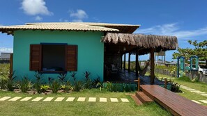 A small, single-story building with a thatched roof porch, surrounded by a well-maintained garden and a wooden deck. - Casa Pindorama com Vista Para o mar em Corumbau (Prado)