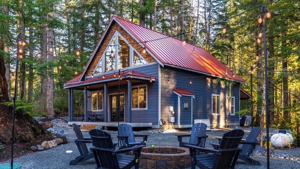 Een chalet met een rood dak, een veranda met vier stoelen en een vuurkuil in het midden.