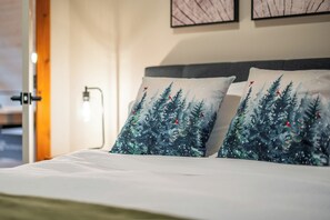 En säng med två kuddar med ett skogsmotiv, ett sängbord med en lampa och en vägg med inramade konstverk.