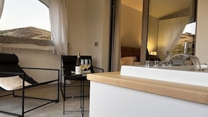A modern room with a bed, a small table with a glass of wine, and a view of a mountainous landscape through a large window.