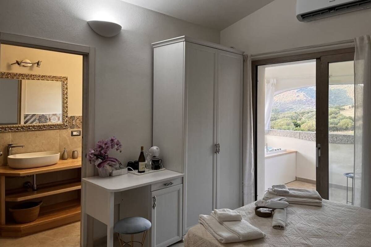 A hotel room with a large bed, a vanity with a sink, a wardrobe, and a balcony with a view of the countryside.