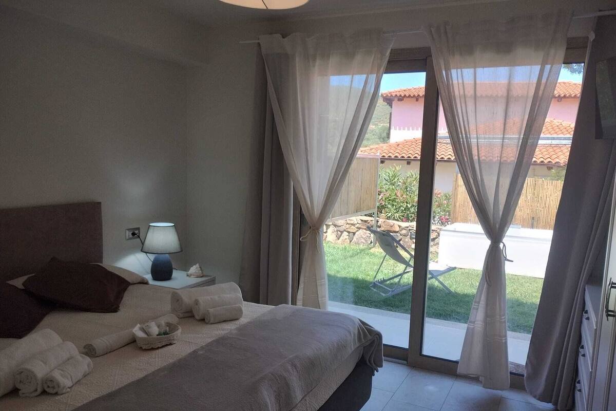 A bedroom with a bed, a nightstand, a lamp, and a view of the outside.