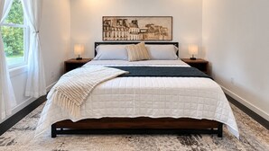 Quarto com cama, duas mesas de cabeceira, ventilador de teto, janela com cortinas e um quadro na parede.