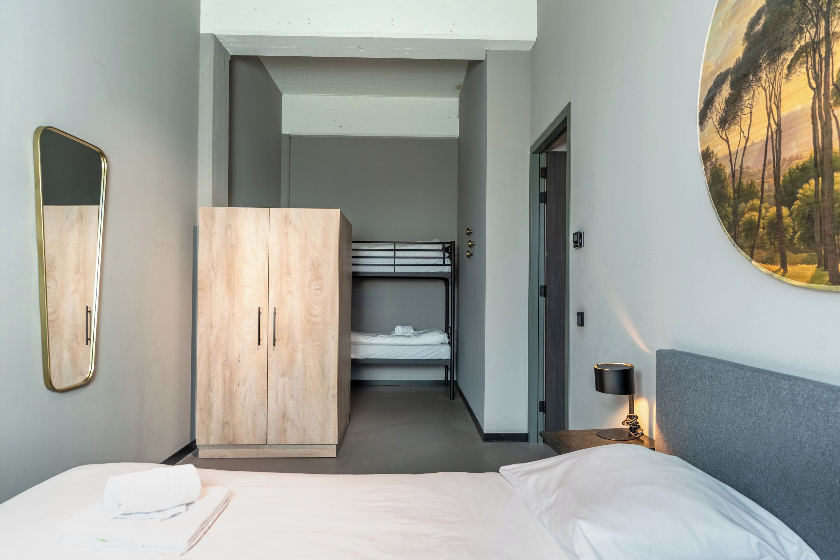 A hotel room with a bed, a wooden wardrobe, a bunk bed, a round painting, a mirror, and a nightstand with a lamp.