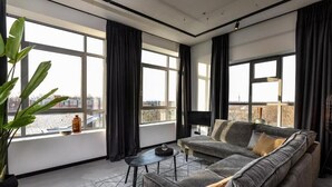 A modern living room with a grey sofa, a small table, and a potted plant. Large windows offer a view of the cityscape.
