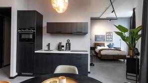 A modern kitchen with dark cabinetry, a built-in oven, and a pendant light. There is a dining area with a table and chairs, and a bedroom with a bed and a large window.