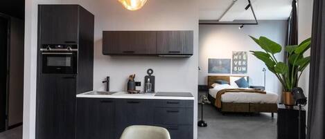 A modern kitchen with dark cabinetry, a built-in oven, and a pendant light. There is a dining area with a table and chairs, and a bedroom with a bed and a large window.