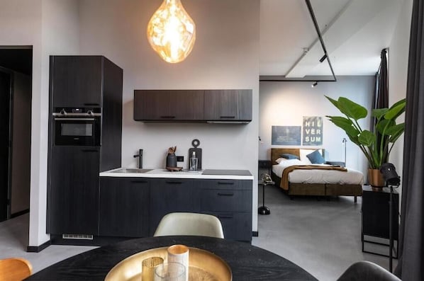 A modern kitchen with dark cabinetry, a built-in oven, and a pendant light. There is a dining area with a table and chairs, and a bedroom with a bed and a large window. - Hilversum City Apartments (Hilversum)