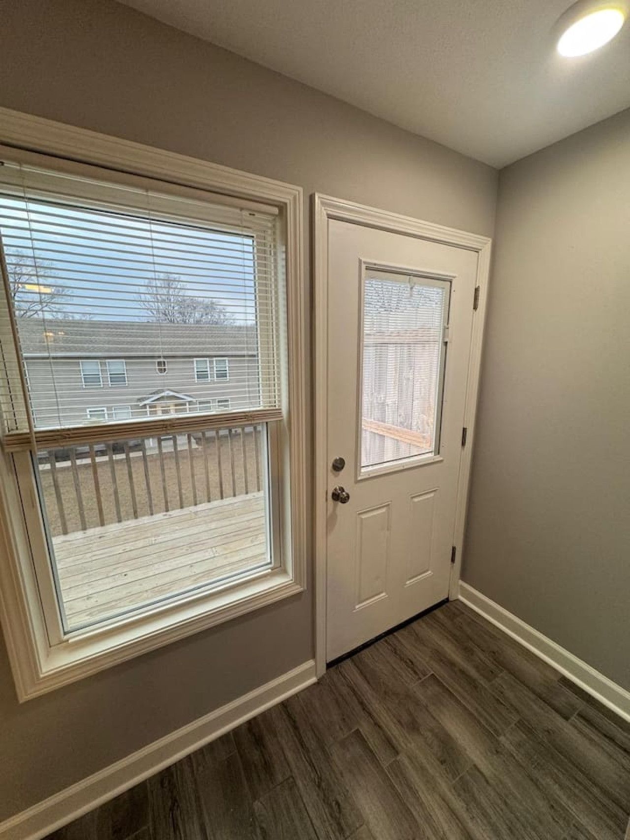 A hallway with a wooden floor, a white door with a window, and a view of a residential area through the window.