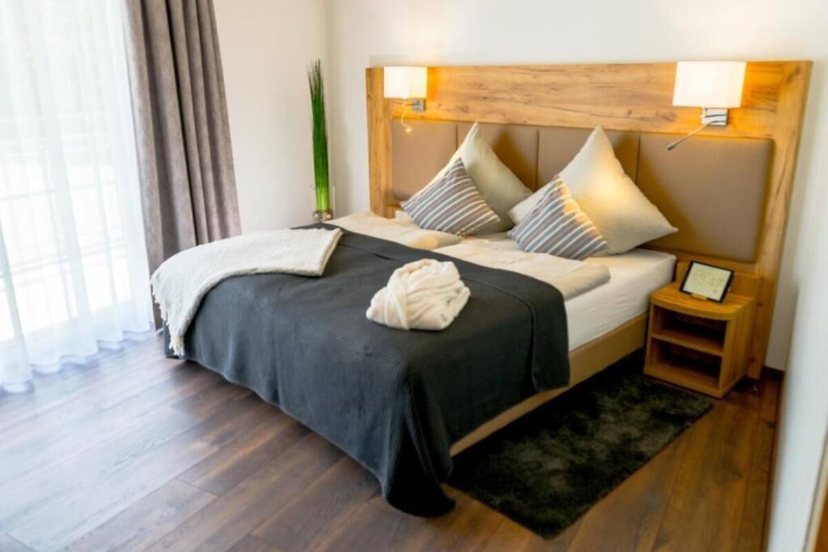 A neatly made bed with a dark bedspread and pillows, a bedside table with a lamp, and a small shelf with a framed photo.