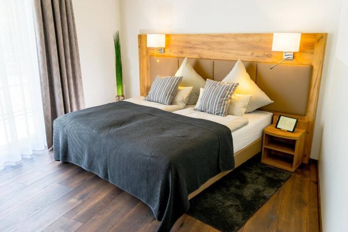 A neatly made bed with a dark bedspread, two bedside lamps, and a small bedside table with a clock.