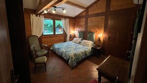 Une chambre à coucher comprenant un lit, une chaise, une table de chevet, une fenêtre avec des rideaux et une structure de plafond en bois.