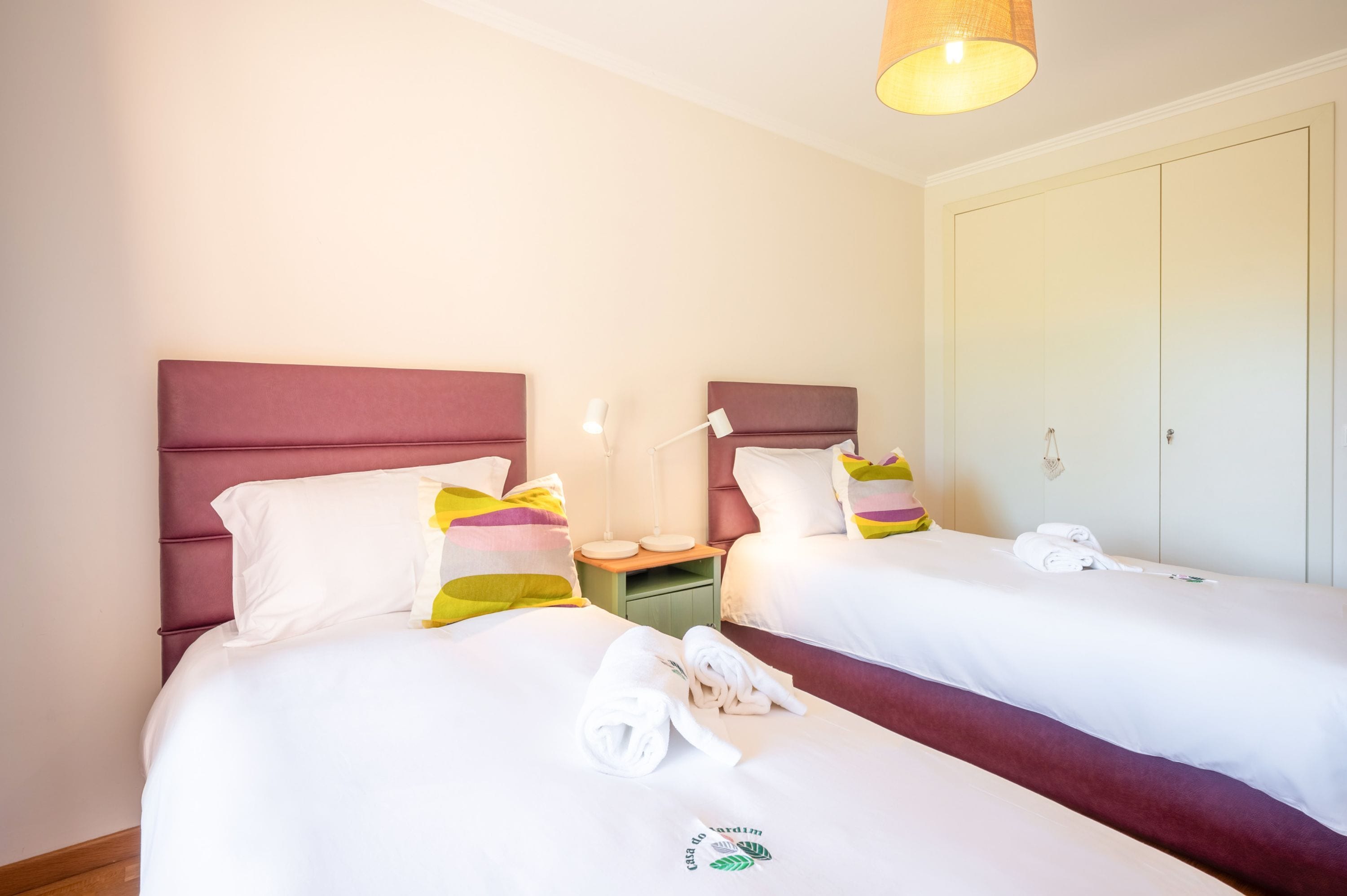 Habitación de hotel con dos camas, cada una con ropa de cama blanca y almohadas coloridas. Entre las camas, una mesita de noche con una lámpara, y un armario con espejo.