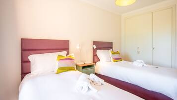 Habitación de hotel con dos camas, cada una con ropa de cama blanca y almohadas coloridas. Entre las camas, una mesita de noche con una lámpara, y un armario con espejo.