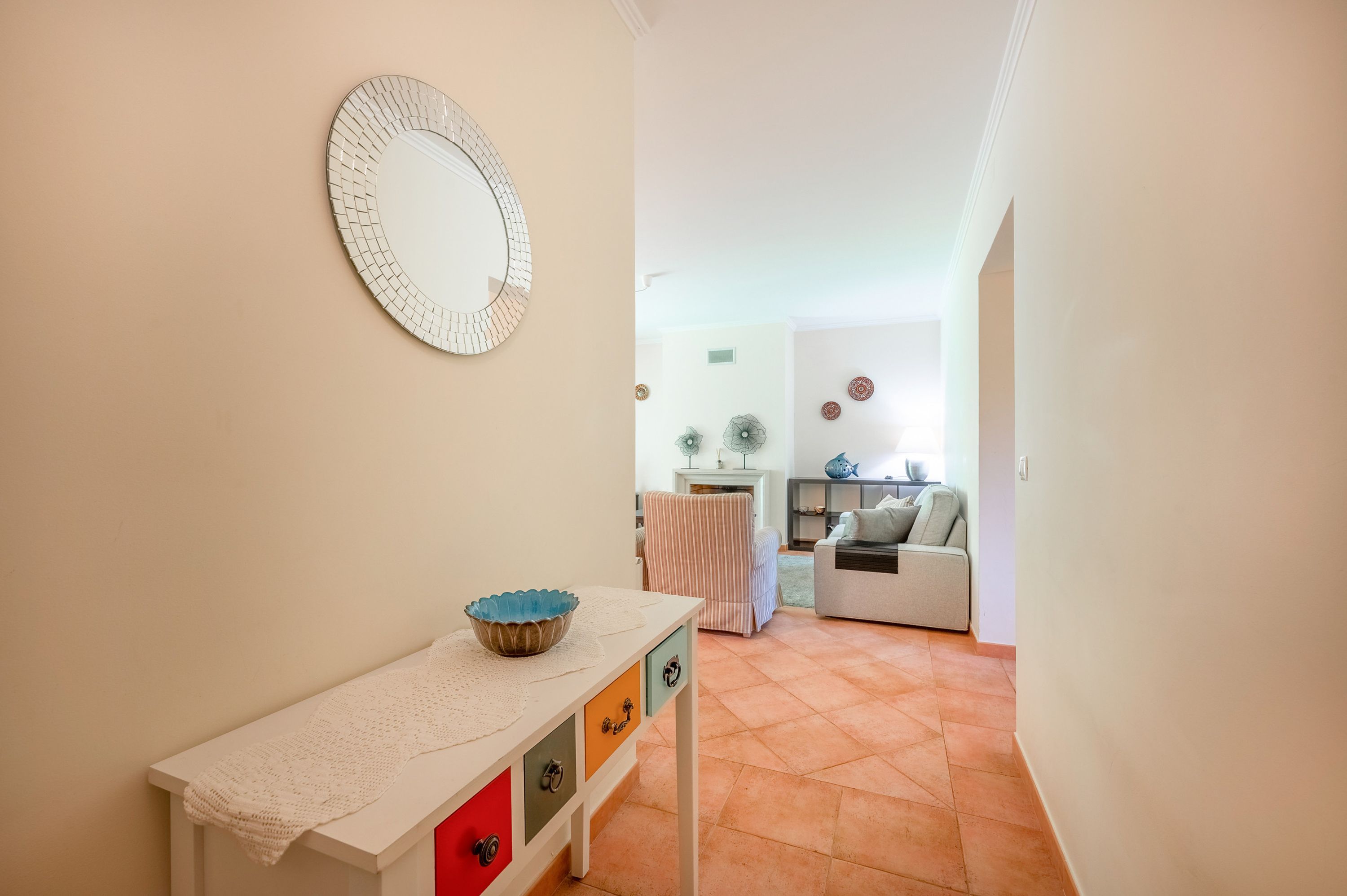 Um corredor leva a uma sala de estar com um sofá branco, uma mesa pequena com uma tigela e um espelho na parede.