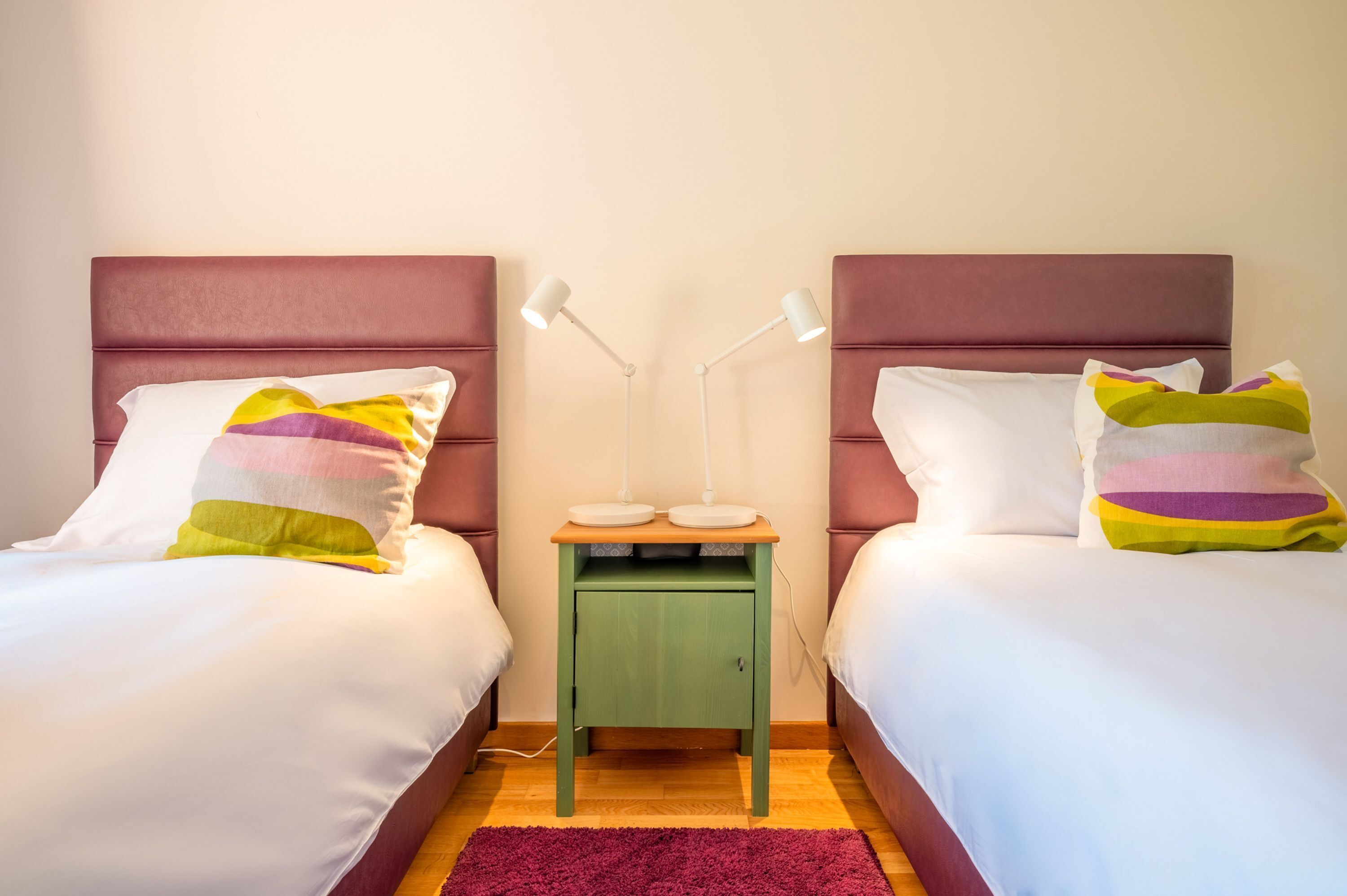 Habitación doble con dos camas, cada una con cabecero, mesita de noche verde pequeña con dos lámparas y alfombra roja.