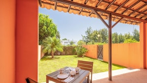 Une terrasse extérieure avec une table dressée pour deux personnes, un banc et, en arrière-plan, une vue sur un jardin et une maison.