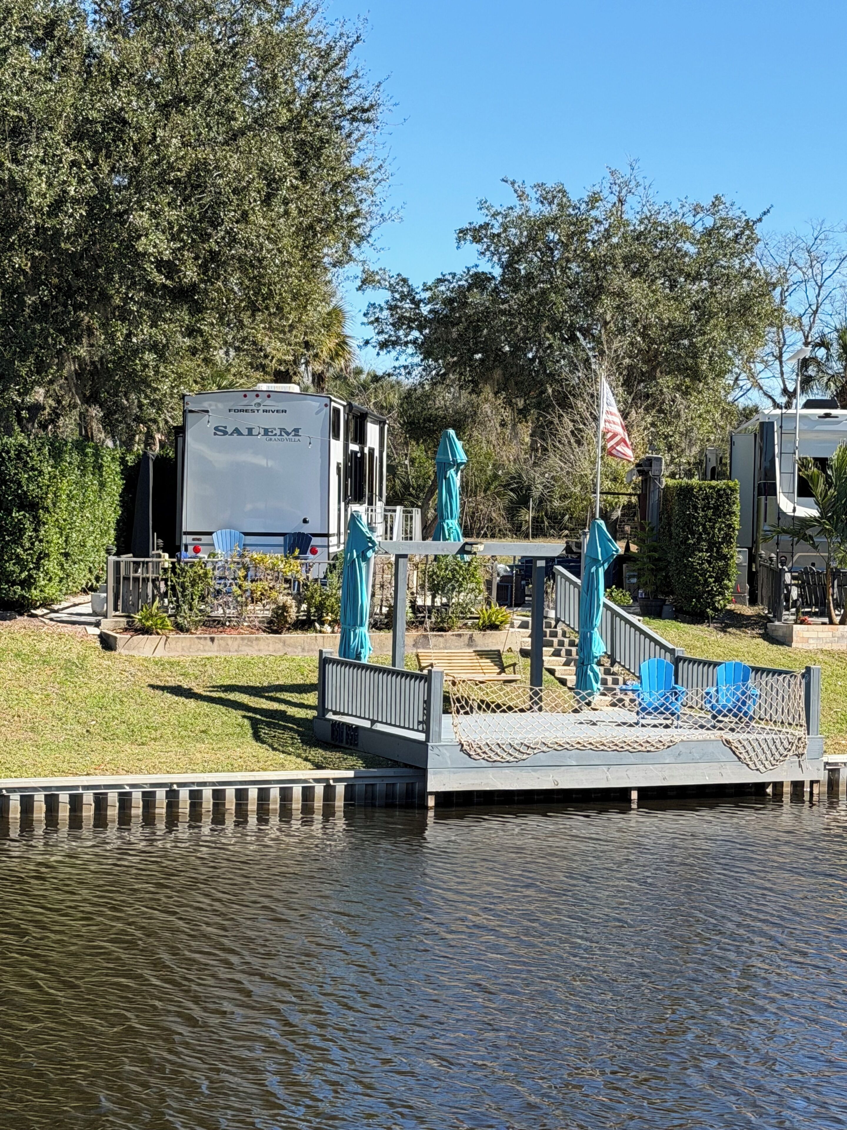 A dock with chairs and a small bridge over water, surrounded by greenery and a large RV.