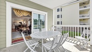 Een overdekt terras met een witte tafel en stoelen, een glazen schuifdeur naar een keuken en een witte balustrade.