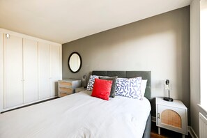 A bedroom with a bed, white wardrobe, bedside table, lamp, and a wall-mounted mirror. - Maida Vale Escape (Greater London)