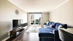 Una sala moderna con televisor de pantalla plana, piso de madera oscura, sofá azul y un ventanal con vista a un edificio.